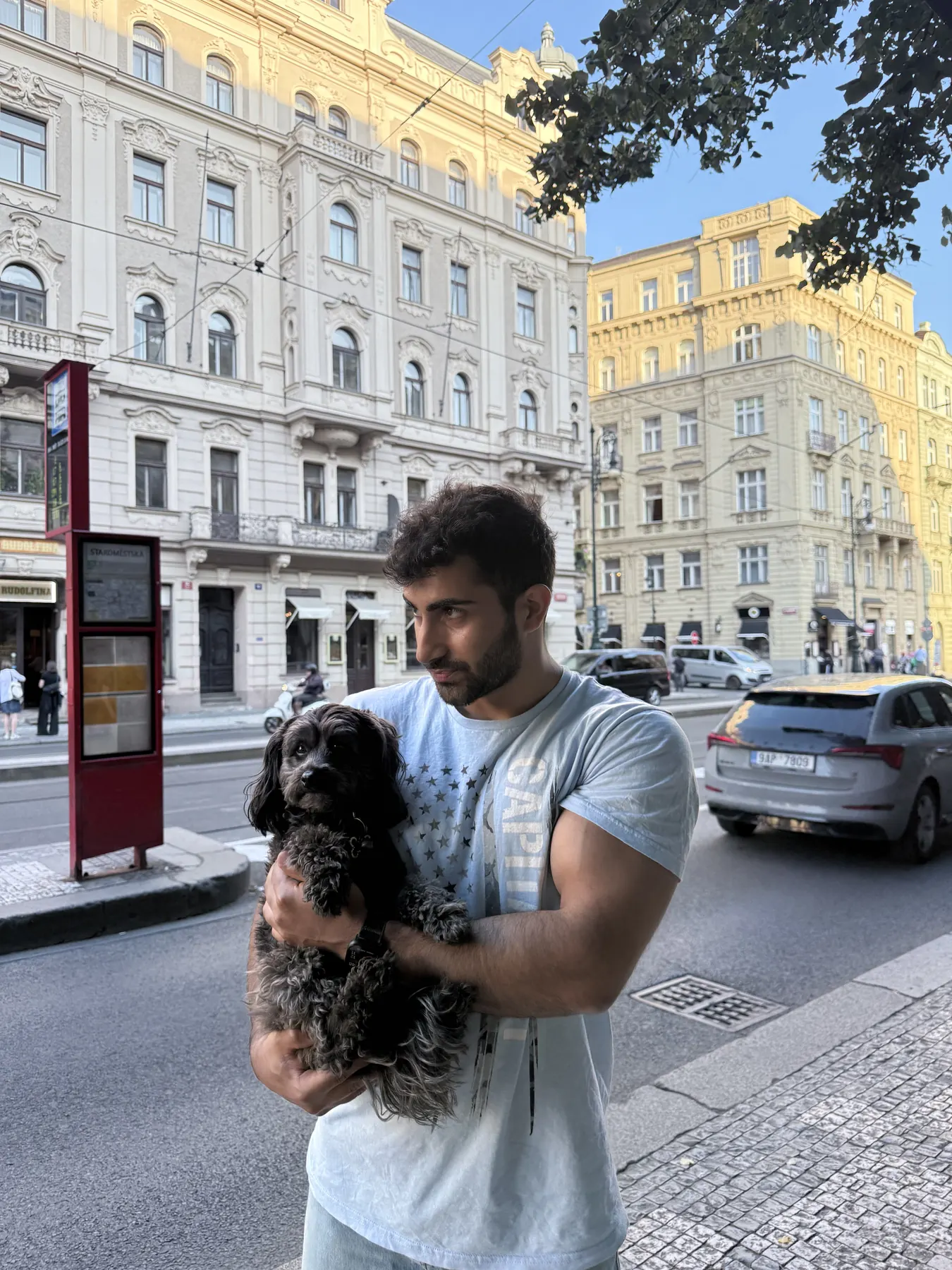 On a Prague street holding a small black dog, period facades behind.